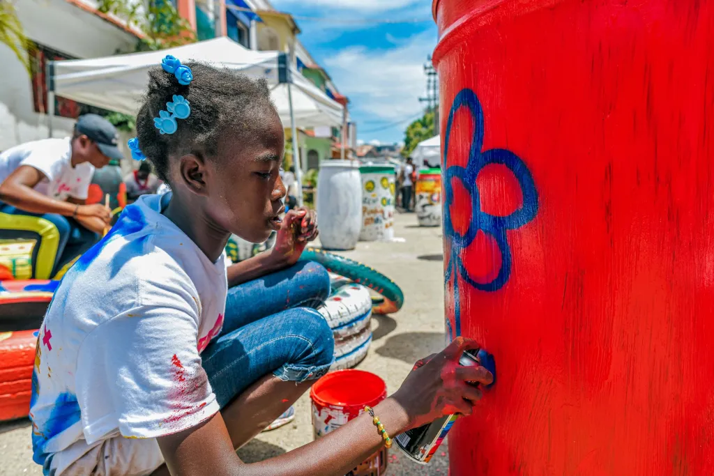 A little girl / A young child sprays a blossom on a trash can. On June 5, PouBèl Ayiti collected locals to clean up debris in Cap-Haitien, place refuse cans in the streets, recycle, and hold a painting competition in honour of World Environment Day. Photo by Oldjy Francois for The Haitian Times