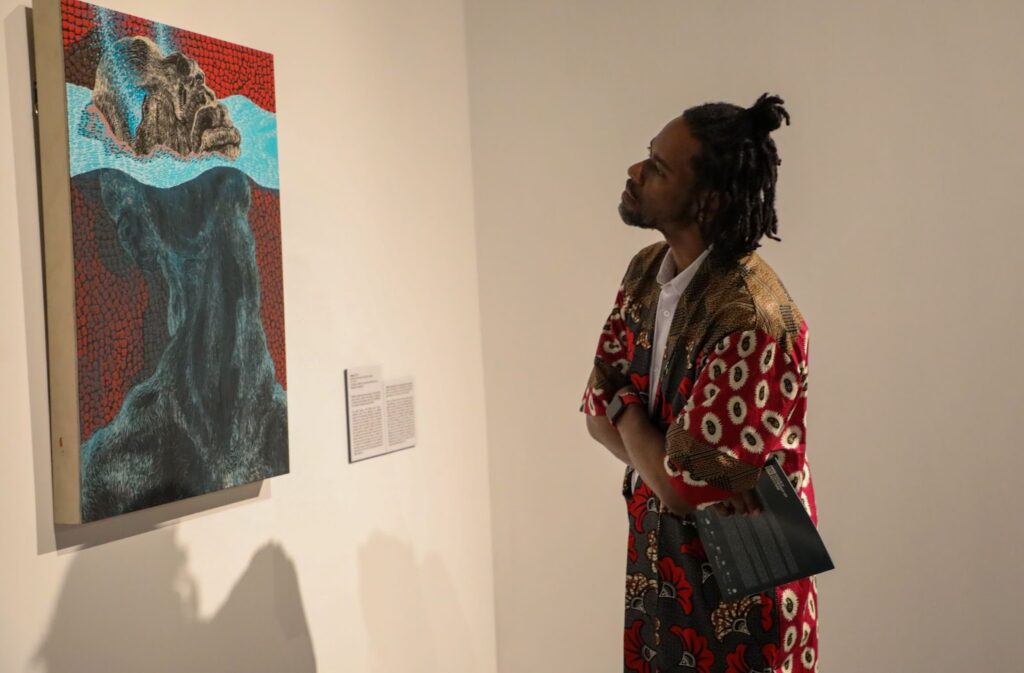 Jerry James / William's friend examines a piece of artwork depicting the migrant experience. "Seeing how he's grown over the year has been fascinating; his career trajectory has been something to behold, especially for a mid-career artist because it usually takes a long time, and his work has been well received."